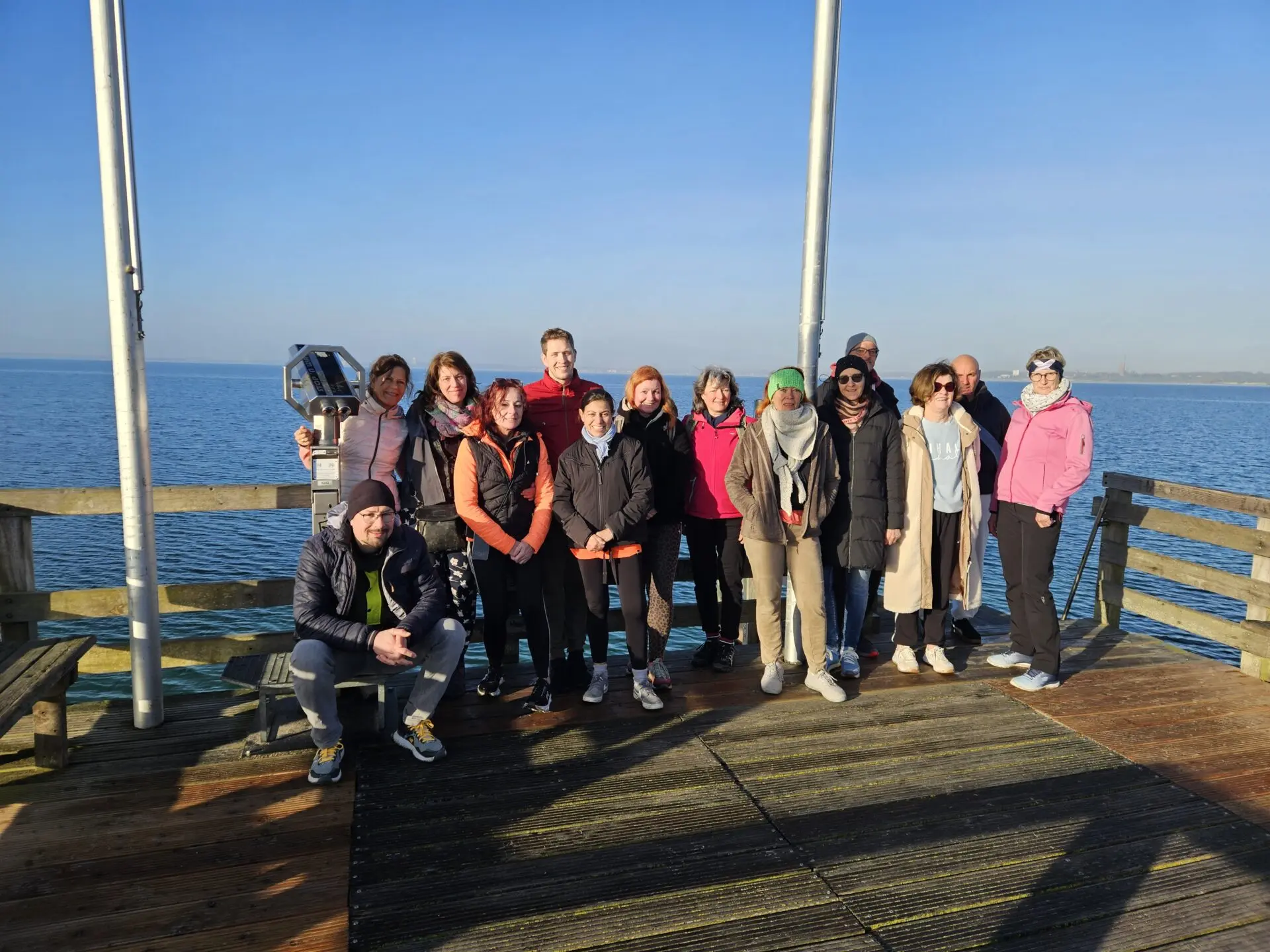 Gemeinsame Gruppe auf einer Holzpromenade am Meer bei klarem, blauem Himmel; Winterjacken, Mützen und Schals vor dem weiten Wasser.